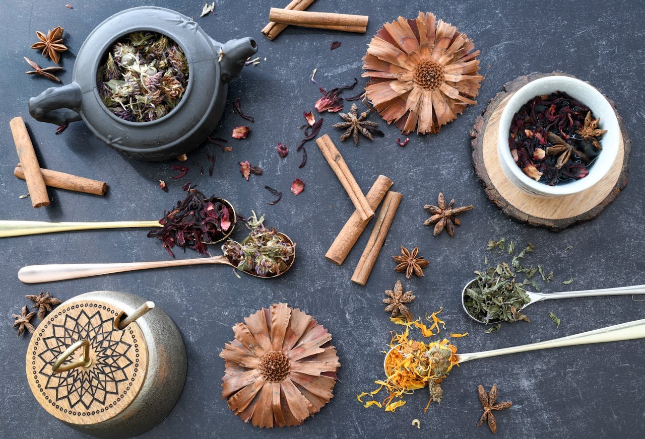 A flat lay of various herbal tea ingredients, spices, and rustic utensils on dark surface.